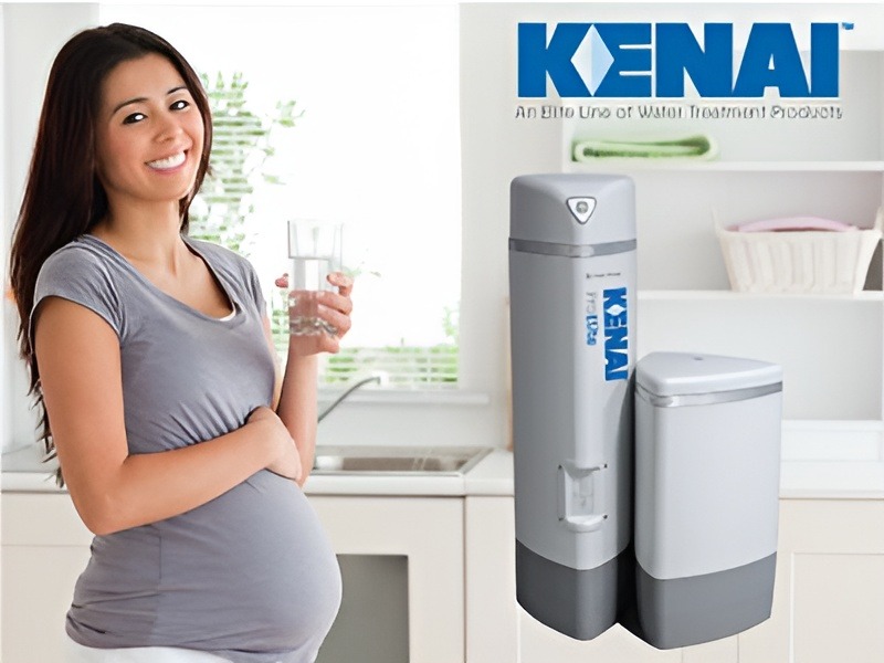 Pregnant woman smiling and holding a glass of water next to a Kenai Elite Line water treatment system in a kitchen.