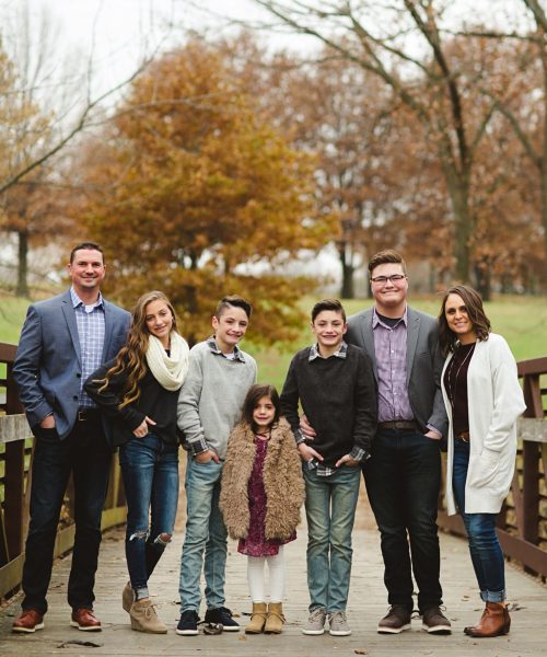 A family of seven stands together on a wooden bridge surrounded by autumn trees. They are dressed in casual and semi-formal attire, with the children in the center and adults on either side, all smiling at the camera.