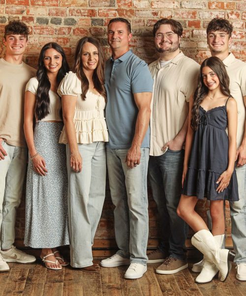 A family of seven stands in front of a brick wall. They are dressed in casual, light-colored clothing and are smiling at the camera. The group includes four young adults, one teenage girl, and two adults.
